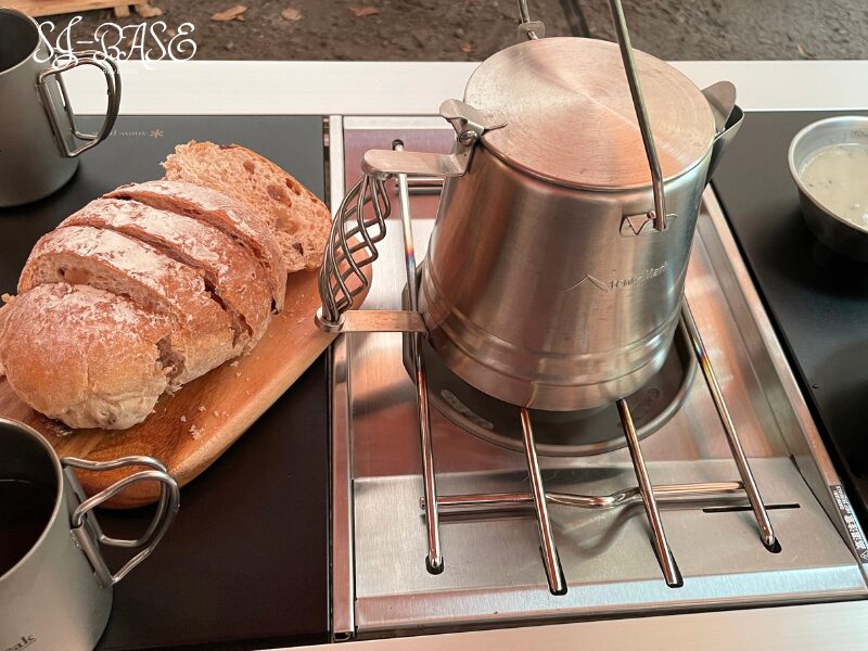 キャンプの朝食準備。ケトルで湯を沸かし、カッティングボードのパンを用意するシーン。