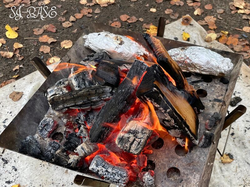 焚き火台で焼き芋を調理中。アルミホイルに包んださつまいもを炭火でじっくり焼いている様子。