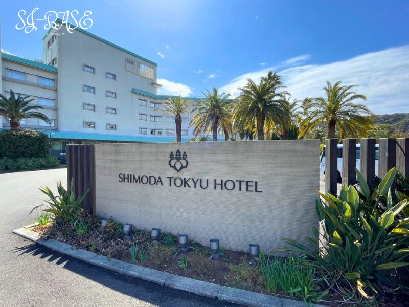 下田東急ホテルのエントランス前にある看板とホテル外観(ヤシの木と青空)